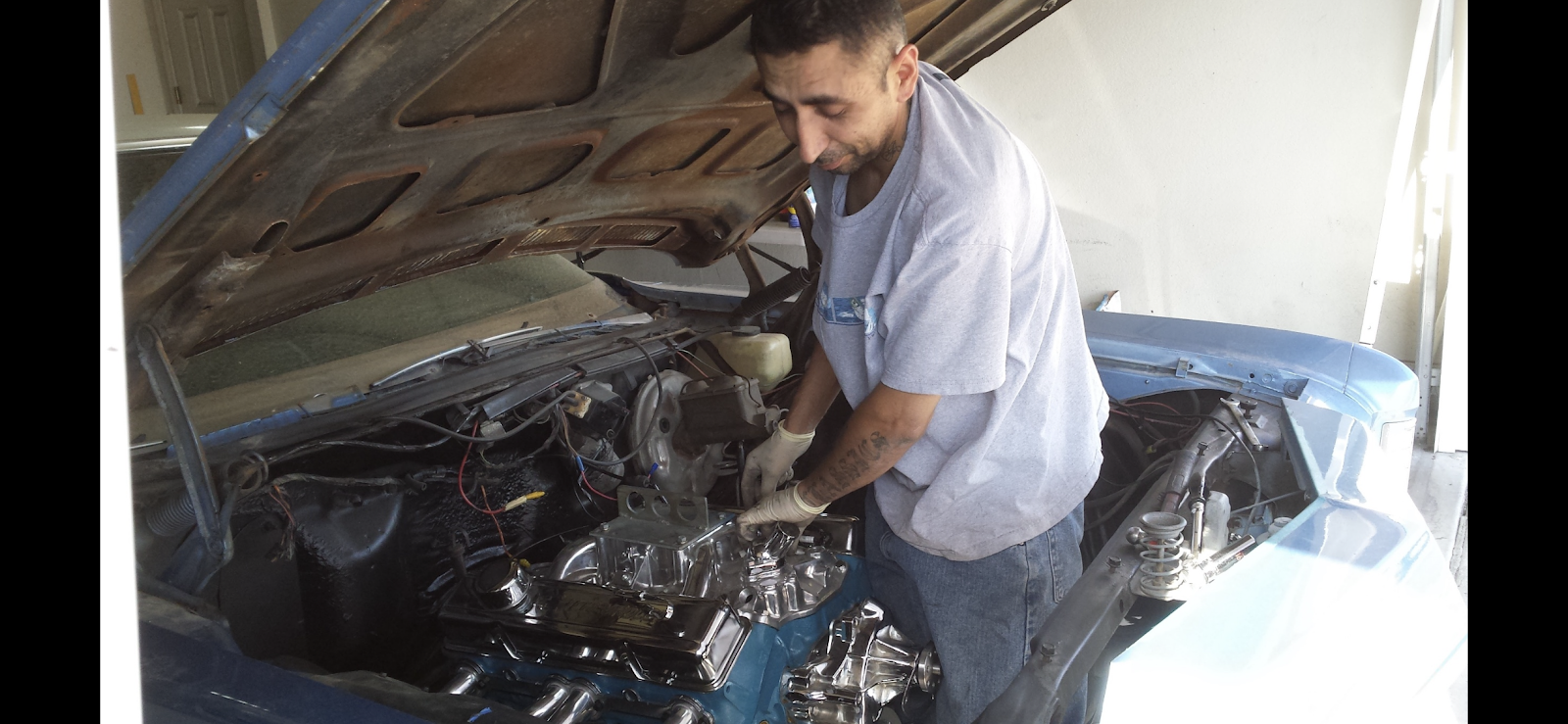 Louie in work gloves reaching into the engine bay of a classic car with chromed valve covers