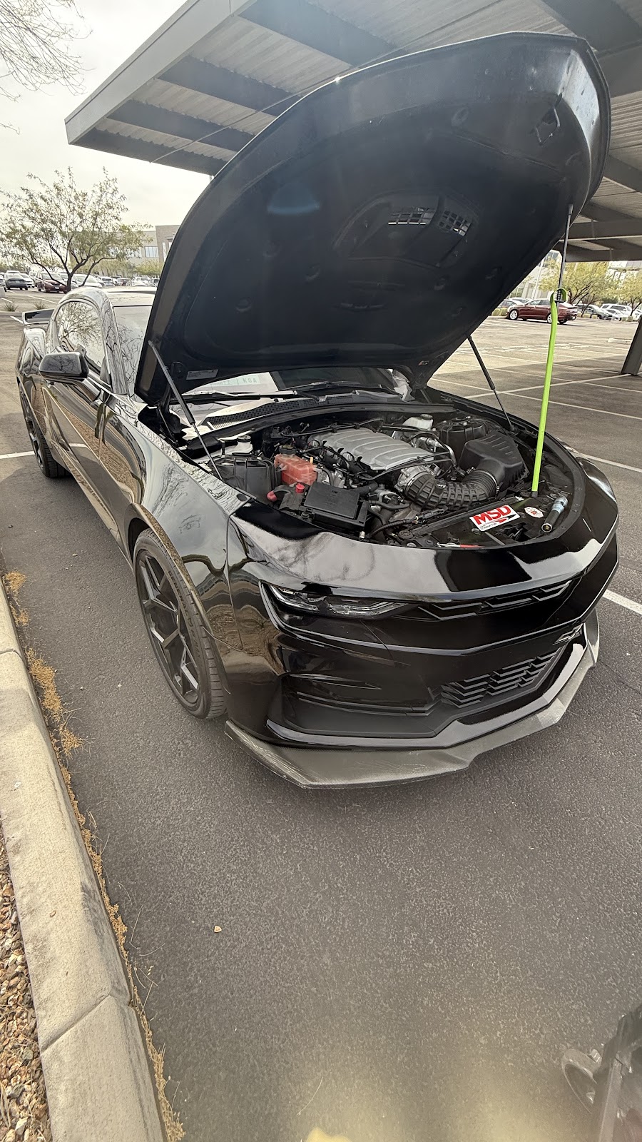 Black Camaro with its hood up under a shade canopy in a parking lot