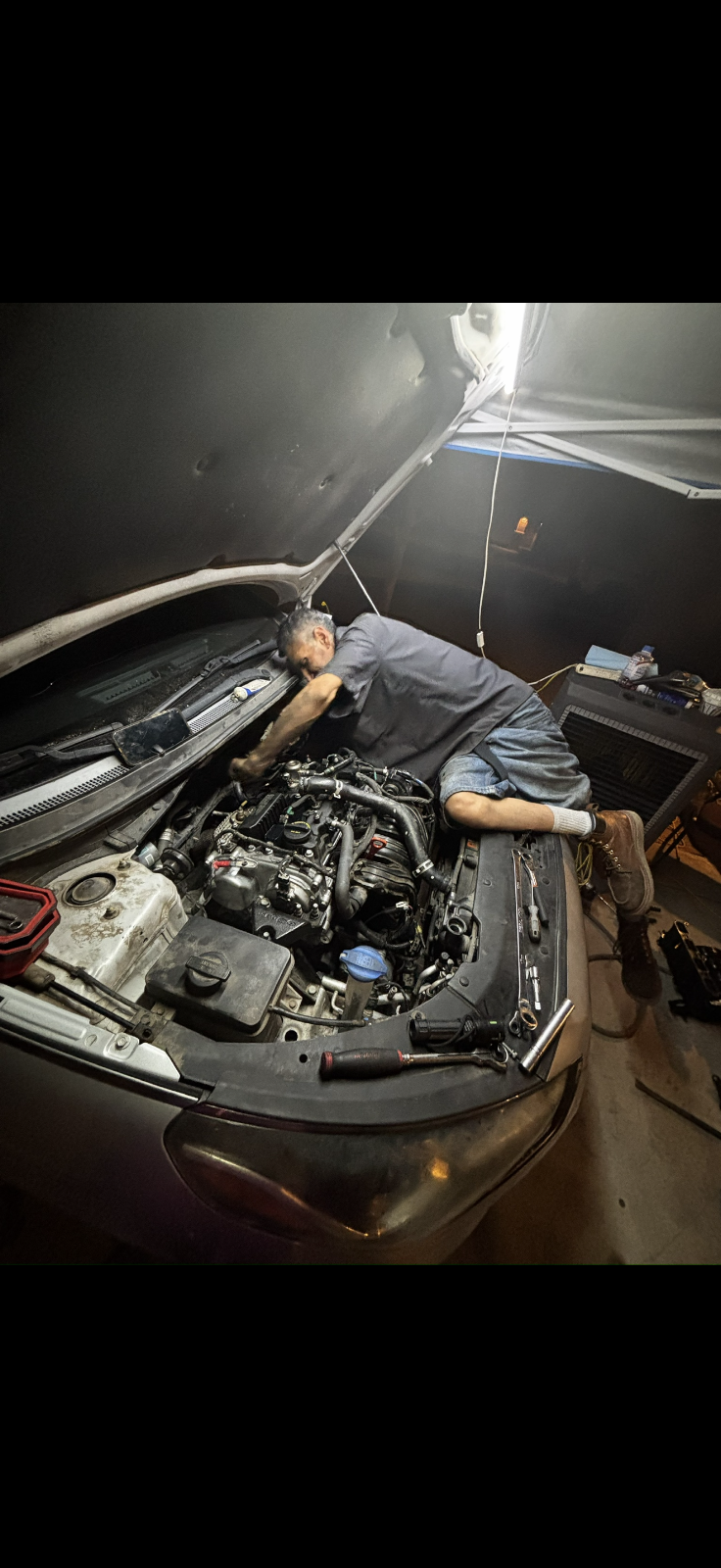 Louie leaning deep into an engine bay at night under a hung work light, sockets lined up along the fender